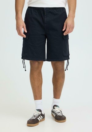 Mann som bærer svarte cargoshorts, hvite sokker og brune Adidas-sneakers, stående mot en ensfarget lys bakgrunn.