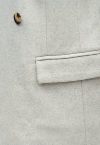 Tissu de manteau en chevrons gris clair avec un bouton rond marron et noir ainsi qu'un rabat de poche passepoilée horizontale.