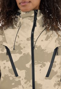 Beige en grijs gestreepte jas met zwarte ritssluitingen, verstelbare capuchon en ventilatiegaten, ontworpen voor buitenactiviteiten.