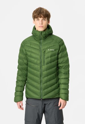 Jonge man draagt een groene doorgestikte gewatteerde jas met capuchon, voorrits en borstzak, gecombineerd met donkergrijze broek tegen een effen achtergrond.
