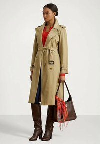 Mulher vestida com um trench coat bege com cinto, top vermelho, jeans escuros, botas castanhas até ao joelho, segurando uma bolsa castanha escura com um lenço de xadrez vermelho.