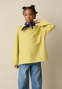 Sárga hosszú ujjú ing laza szabással, magas derekú kék farmernadrággal párosítva. Egy tengerészkék bandana részletként a nyak körül.