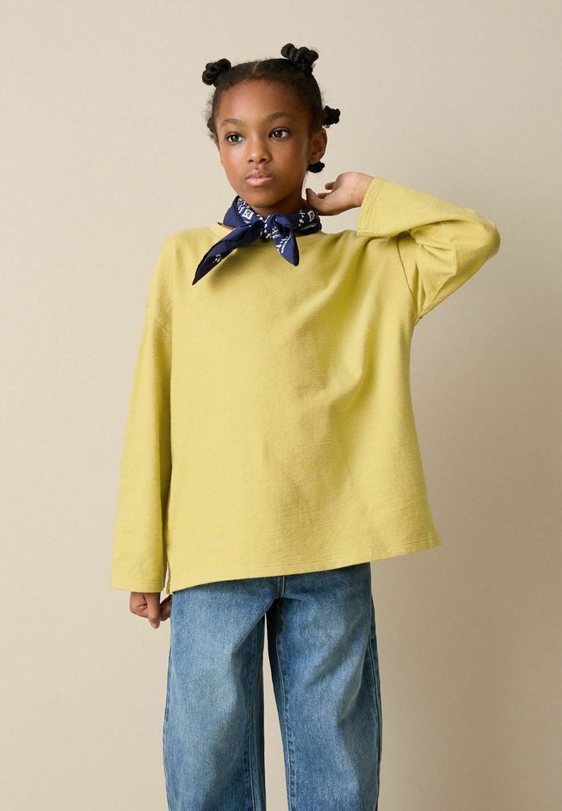 Sárga hosszú ujjú ing laza szabással, magas derekú kék farmernadrággal párosítva. Egy tengerészkék bandana részletként a nyak körül.