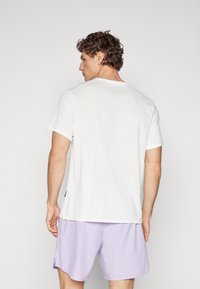 Hátsó nézet egy rövid ujjú, fehér pólóról, amely texturált mintával és kerek nyakkivágással rendelkezik, világoslila rövidnadrággal párosítva.