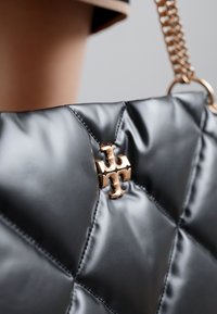 Bolsa de mão preta, acolchoada e com acabamento brilhante, apresentando um detalhe em logótipo dourado e alça em corrente. Superfície texturizada com padrão de diamante.