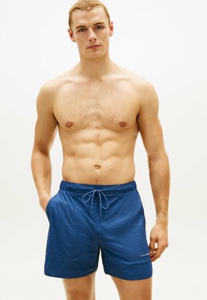 Oberkörperfreier Mann mit kurzen Haaren, trägt blaue Badeshorts mit Kordelzug, die Hände in den Taschen, neutraler Hintergrund.