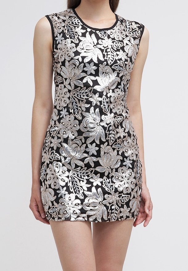 Mini-robe noire sans manches avec broderie florale argentée portée par une personne debout devant un fond clair uni.