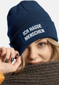 Navy knitted beanie with a rectangular patch displaying the text “ICH HASSE MENSCHEN” in white. Textured fabric with a snug fit.