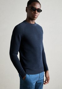 Marineblauer Strickpullover mit einem strukturierten Muster, Rundhalsausschnitt und gerippten Manschetten, kombiniert mit hellblauen Hosen. Flach gelegte Hände in den Taschen.