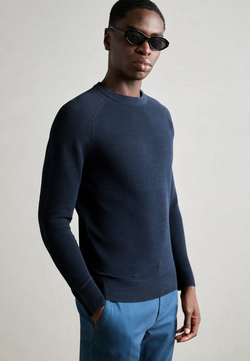 Marineblauer Strickpullover mit einem strukturierten Muster, Rundhalsausschnitt und gerippten Manschetten, kombiniert mit hellblauen Hosen. Flach gelegte Hände in den Taschen.