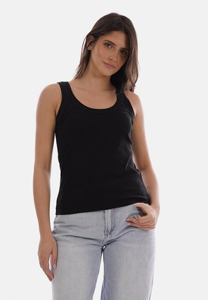 Donna con capelli castani lisci, che indossa una canotta nera senza maniche e jeans azzurri, in piedi contro uno sfondo chiaro e uniforme.