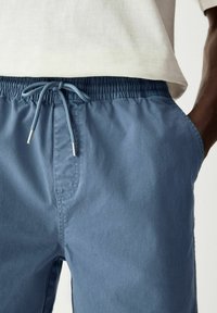 Pantalones cortos azul claro con cordón ajustable en la cintura, bolsillos frontales y una textura suave. Los cordones tienen puntas de metal.