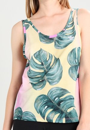 Vrouw in een mouwloos Adidas-top met grote groene tropische bladeren op een pastelgele en -roze achtergrond.