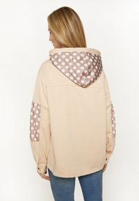 Beige oversized jacka med blommigt mönster på huvan och ärmarna, med en mjuk textur och knapppdetaljer.