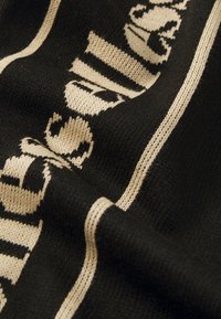 Μαύρος πλεκτός ύφασμα με μπεζ σχέδιο που περιλαμβάνει κείμενα με υφή και οριζόντιες ρίγες, που δείχνει τον μαλακό και ευέλικτο σχεδιασμό του νήματος.