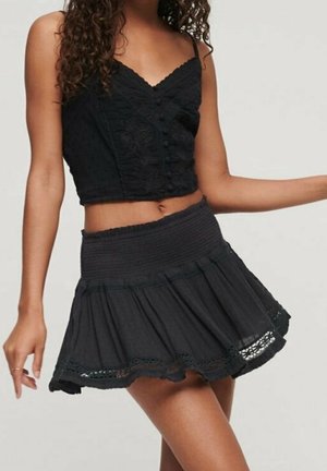 Femme portant un crop top en dentelle noire avec des boutons et une jupe noire taille haute avec bordure en dentelle, debout avec un bras tendu.