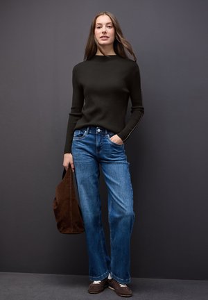 Dunkel gestreifter Pullover, blaue weitgeschnittene Jeans, braune Wildledertasche. Das Modell steht vor einem grauen Hintergrund und präsentiert ein lässiges Outfit.