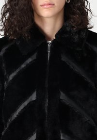 Jachetă din blană artificială neagră cu un model în zigzag, închidere cu fermoar și guler. Textură moale și finisaj lucios care îmbunătățește detaliile de design.