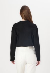 Czarny sweter z prążkowanego materiału, z wysokim dekoltem i długimi rękawami, w zestawie z białymi dżinsami. Widoczne detale przeszycia i luźny krój.