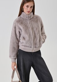Giacchino grigio in pelliccia sintetica, corto, con colletto alto, chiusura con zip e polsini a coste. Abbinato a jeans scuri a vita alta e una borsa neutra.