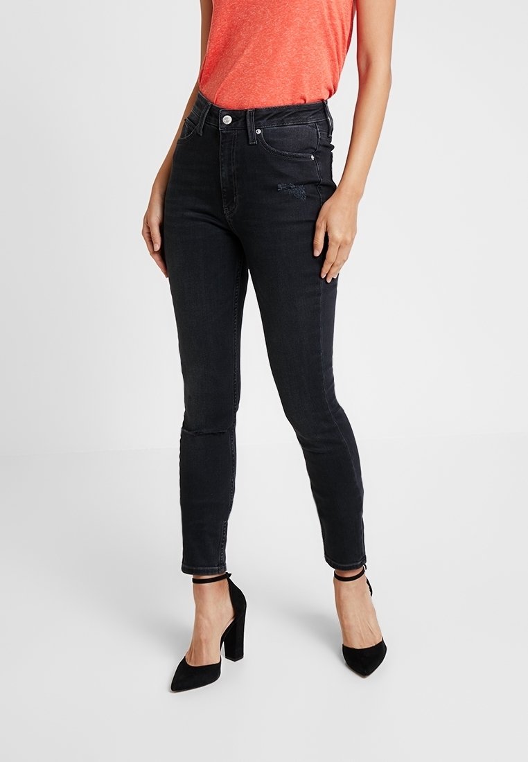 Jeans pitillo negros de tiro alto en tela vaquera con ligero desgaste en la parte delantera, combinados con una camiseta roja y tacones negros.