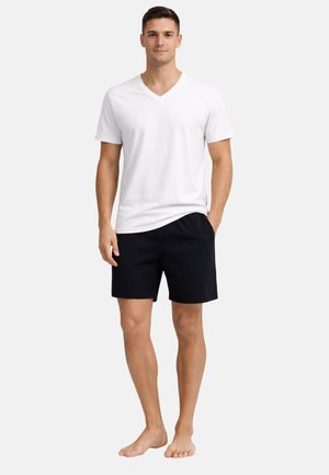 Muž stojící naboso, oblečený v bílem tričku s V-výstřihem a černých šortkách, s jednou rukou v kapse, neutrální pozadí.