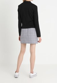 Musta pitkähihainen takki, jossa on räätälöity istuvuus, yhdistetty harmaaseen ja valkoiseen ruutuhameeseen. Mallilla on valkoiset lenkkarit.