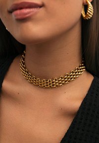 Złota metalowa choker z warstwowym wzorem łańcucha. Posiada gładką, odbijającą powierzchnię i ściśle przylega do szyi. Małe kolczyki w kształcie kółek dopełniają całość.