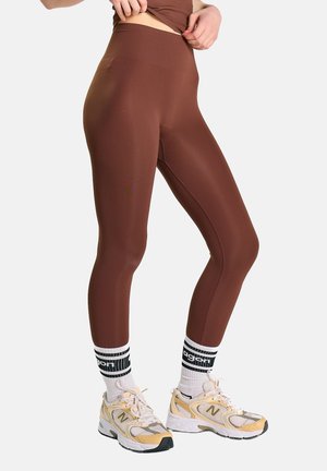 Personne portant des leggings marron taille haute, des chaussettes blanches avec des rayures noires et du texte, et des baskets New Balance beige, debout sur un fond blanc.