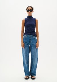 Canottiera a costine blu navy senza maniche, jeans a vita alta blu chiaro a gamba larga e sandali neri flati. Il modello è in piedi contro uno sfondo bianco.