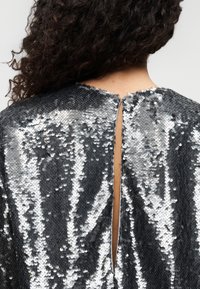 Το ύφασμα με ασημένιες πούλιες διαθέτει υφή με υψηλή γυαλάδα και σχεδιασμό στην πλάτη με άνοιγμα σε σχήμα κλειδιού, παρουσιάζοντας λαιμόκοψη με στρογγυλεμένο και ομαλό περίγραμμα και λαμπερές λεπτομέρειες.