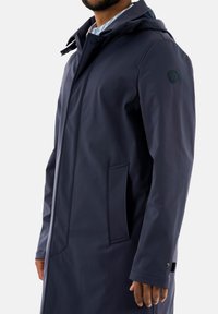 Abrigo impermeable azul marino con capucha, con cierre de cremallera, bolsillos laterales y un logo en la manga. Textura suave y diseño moderno.
