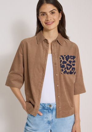 Femme souriante aux longs cheveux foncés portant une chemise marron à boutons avec une poche imprimée léopard par-dessus un haut blanc et un jean bleu clair.
