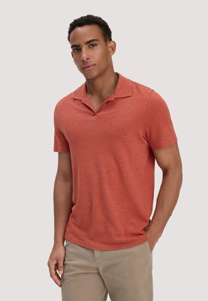 Man draagt een roestkleurig poloshirt met korte mouwen en beige broek, staand met één hand in de zak tegen een effen achtergrond.