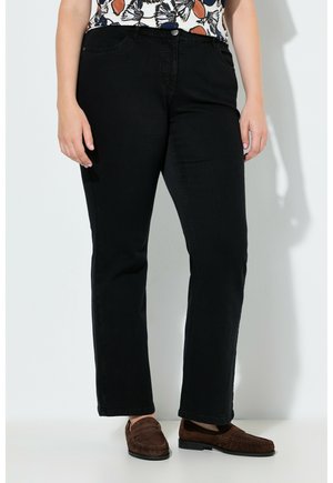 Person iført sorte bukser med lige ben, en mønstret skjorte og brune loafers, der står op ad en hvid væg.