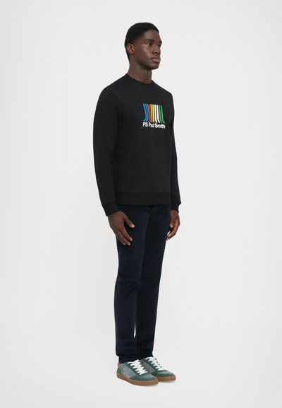 Mann som står iført svart hettegenser med fargerik stripet logo og tekst, mørke bukser og grønne joggesko på hvit bakgrunn.