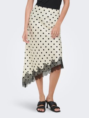 Femme portant une jupe midi crème à pois noirs avec un ourlet en dentelle noire festonné, assortie à des sandales à talons noires et un haut noir.