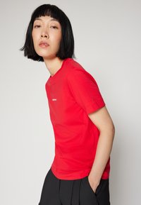 Red cotton t-shirt with a crew neckline, short sleeves, and a small logo centered on the chest. Paired with black trousers.