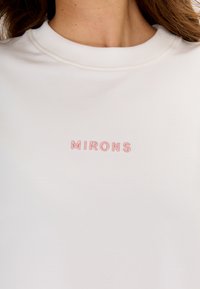 Witte sweatshirt met een geribbelde ronde hals. Heeft subtiele roze geborduurde tekst "MIRONS" op de voorkant. Gladde textuur.