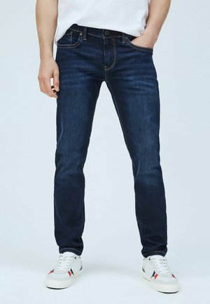 Hombre con vaqueros azul oscuro de corte ajustado, camiseta blanca y zapatillas blancas con detalles negros y rojos, de pie con una mano en el bolsillo.