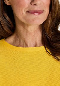 Frau mit braunen Haaren, die ein gelbes Strickoberteil trägt, zeigt den unteren Teil des Gesichts und den Hals mit einem subtilen Lächeln und glänzenden Lippen.