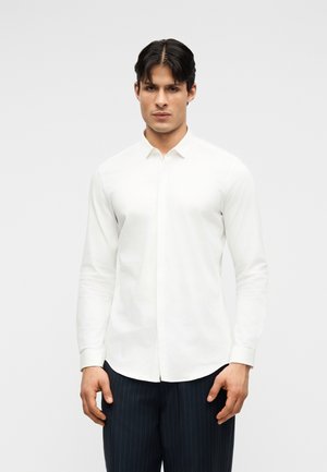 Άνδρας με σκούρα μαλλιά, φορώντας εφαρμοστό λευκό πουκάμισο με μακριά μανίκια και μαύρο παντελόνι με ρίγες, στέκεται μπροστά σε απλό φόντο.