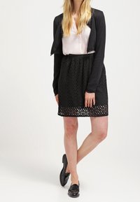 Schwarze, kurze Strickjacke, hellrosa Bluse und knielanger schwarzer Rock mit geometrischen Ausschnittmustern. Schwarze Loafers runden das Outfit ab.