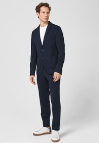 Marineblauer Anzug mit einem maßgeschneiderten Blazer und zwei Taschen, kombiniert mit passenden Hosen und weißen Sneakers, die ein modernes Design präsentieren.