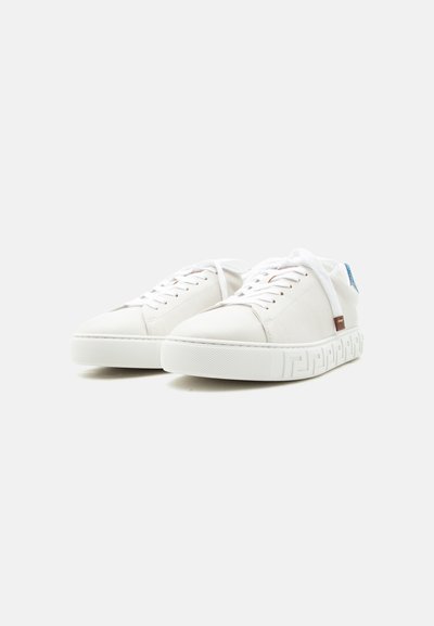 Paire de baskets blanches basses avec lacets, semelles texturées et petits accents bleus et bruns, posées sur un fond clair uni.