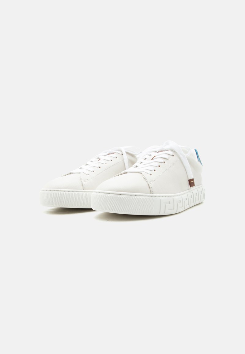 Paire de baskets blanches basses avec lacets, semelles texturées et petits accents bleus et bruns, posées sur un fond clair uni.