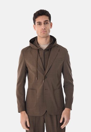 Uomo con abito marrone e felpa con cappuccio con cerniera indossata sotto, in piedi davanti a uno sfondo chiaro e uniforme.