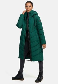 Cappotto puffer verde, design trapuntato con cappuccio, chiusura con zip, tasche laterali e dettagli neri. Abbinato a jeans scuri e stivaletti neri con lacci.