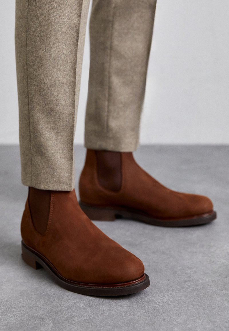 Brown suede Chelsea boots with elastic side panels and a round toe, featuring a contrasting dark brown leather sole. Trousers in light beige.