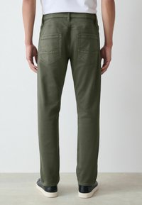 Oliivrohelised sirged püksid, tagaküljel kaanepocketid ja tagasihoidlikud õmbluse detailid. Kandmisel mustade tossudega heledal taustal.